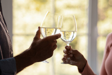 Cropped young husband and wife clink glasses with wine on window background