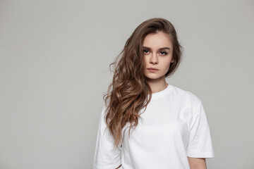 Female portrait of a beautiful girl with a hairstyle in a white T-shirt in the studio looks at the camera. Serious woman on gray background