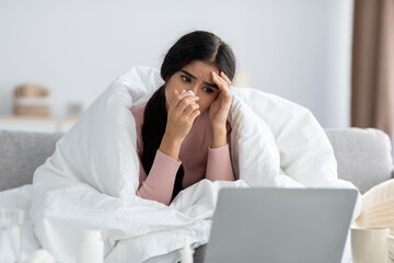Sad young indian woman wrapped in blanket, suffering from flu sits on sofa wipe nose looks at laptop