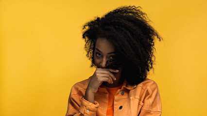 young and shy african american woman covering face with curly hair isolated on yellow.