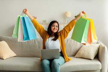 Overjoyed arab lady shopping online, sitting on sofa with laptop, lifting gift bags above her head in excitement