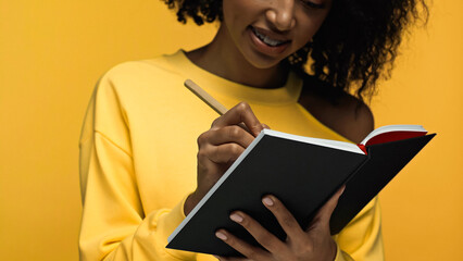 cropped view of happy african american woman with braces writing in notebook isolated on yellow.