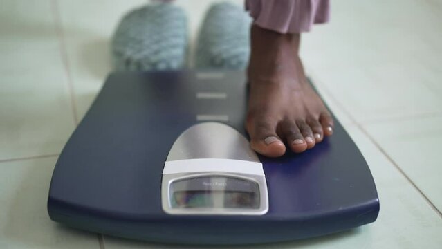 Close-up bathroom scales with barefoot African American female feet stepping on device. Unrecognizable slim young woman measuring body weight indoors at home in the morning