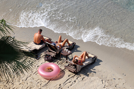 Man and two young women in bikini sunbathing on a sandy beach, top view. People with smartphones lying in deck chairs, vacation on a sea resort