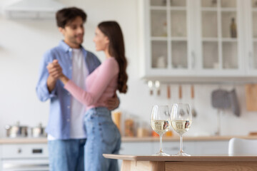 Cheerful young european man and lady dancing in kitchen interior, blurred, focus on two glasses with wine