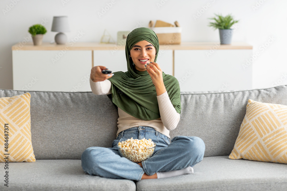 Wall mural Young Arab woman in hijab watching TV and eating popcorn on couch at home