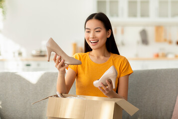 Happy asian woman received package, unpacking cardboard box, holding and looking at high heel...