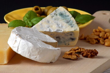 Three cheeses - camembert brie, camembert blue and parmesan cheese stacked on top of each other next to honey, olives on a plate and basil