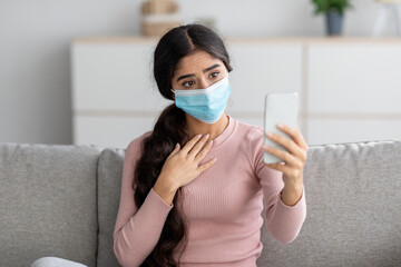 Sad young indian lady in protective mask on sofa makes selfie on smartphone or video call in living...