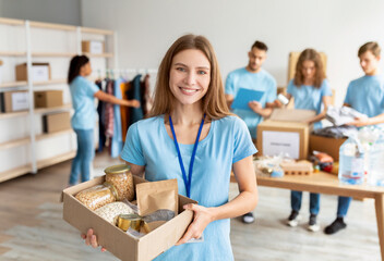 Positive female volunteer in blue uniform holding cardboard box with food donation, working in...