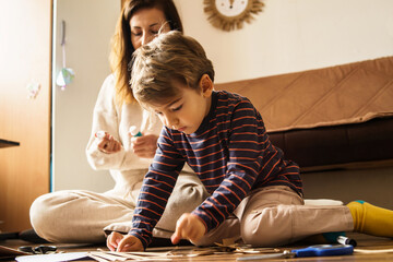 Mother and son playing at home having fun in room cutting paper with scissors making craft and art...