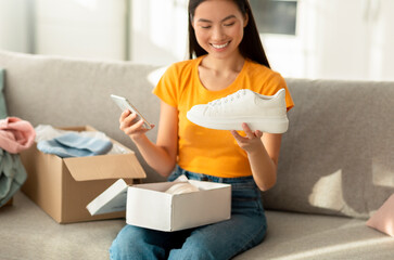 Positive asian woman unpacking parcel with new sports shoes and holding smartphone, writing online store feedback - obrazy, fototapety, plakaty