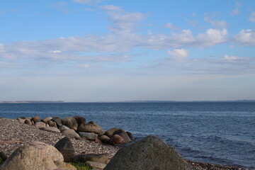 Fototapeta na wymiar Steinstrand an der Nordsee. Die Küste entlang des Ozeans ist sandig. Die Gegend ist felsig, grün. Der Himmel verschmilzt mit dem Wasser am Horizont. Vom Wind getragene weiße Wolken ziehen schnell.