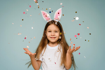Beautiful happy little girl with bunny ears with confetti on a blue background celebrates easter.