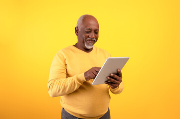 Positive elderly black man using digital tablet for freelancing job or studies on orange studio background