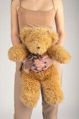 Woman and child, fashion concept. Slim and beautiful woman with beige trousers and t shirt without sleeves holding fluffy brown bear toy in her hands. Studio shot in grey background