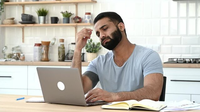 Tired exhausted indian man, freelancer, designer, IT specialist, working remotely at home, overworked by working in a laptop, finished work, closed laptop, needs a break, looks to the side