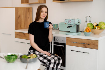 Young beautiful vegetarian girl dressed in pajamas eating fruits and vegetables for breakfast at home in the kitchen