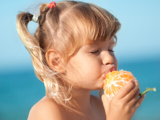 Little girl eats a very tasty mandarin on the beach. Her eyes are closed. Sea background.	