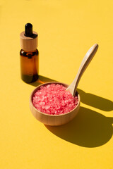 Wooden bowl with a spoon filled with pink bath sea salt. Beauty treatment for spa and wellness on yellow background. Skin care natural cosmetic concept for body care. Dropper bottle with essential oil