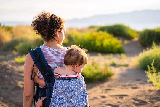 Back of a mother walking carrying a baby on her back in a baby carrier outdoors