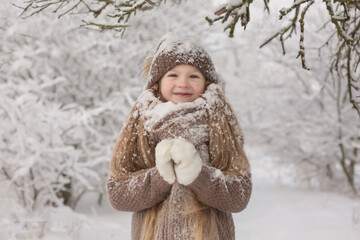 a beautiful long-haired little blonde girl in brown knitted things a hat and a scarf is standing in a snowy winter park