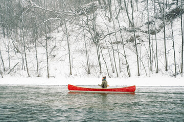 A man riding a red Canadian canoe on the snowy winter river.