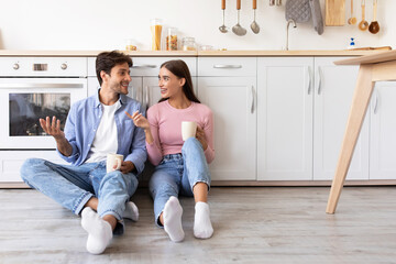 Smiling millennial european woman and man with cups of drink, sitting on floor, talk in kitchen...