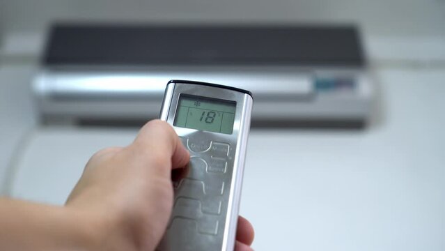 man's hand with remote control is aimed at air conditioner. By moving your finger and pressing button, numbers on display change, raising temperature and cooling it. Air conditioning and heating.