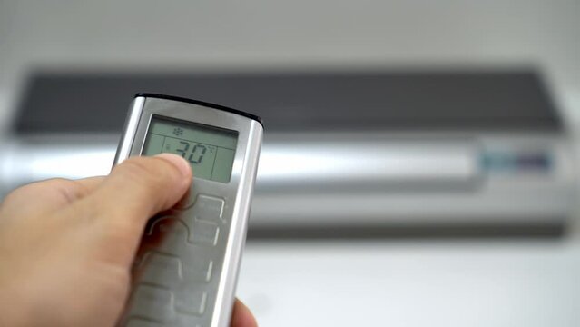 man's hand with remote control is aimed at air conditioner. By moving your finger and pressing button, numbers on display change, raising temperature and cooling it. Air conditioning and heating.