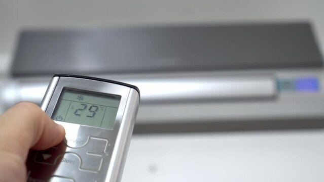 man's hand with remote control is aimed at air conditioner. By moving your finger and pressing button, numbers on display change, raising temperature and cooling it. Air conditioning and heating.