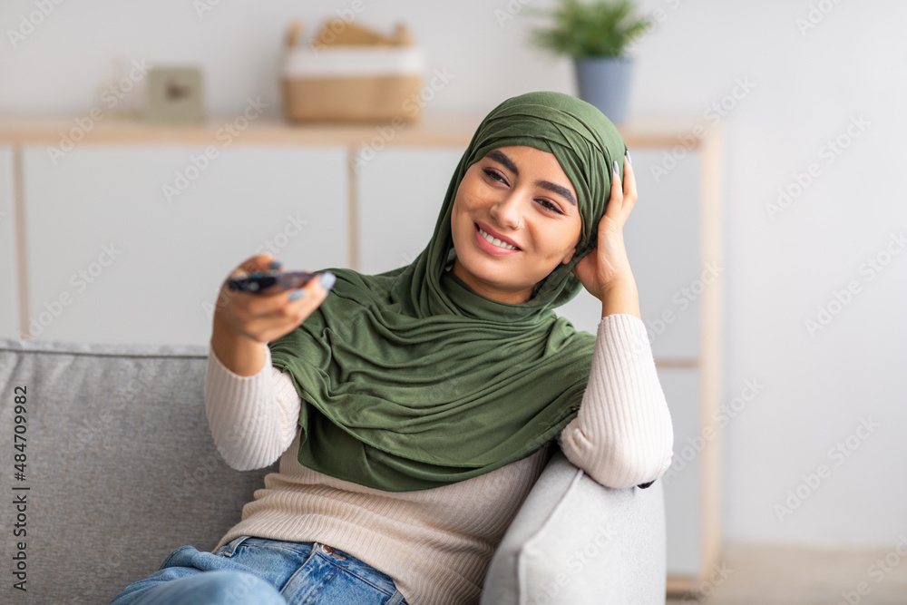 Wall mural young arab woman in muslim hijab holding remote control, watching tv on sofa at home