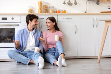 Cheerful smiling young wife and husband with cups of hot drink sitting on floor and talking at...