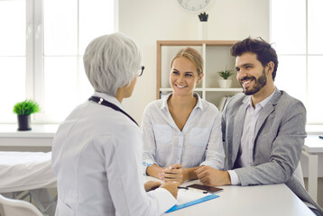 Happy young married couple in hospital consults with experienced doctor about planning pregnancy. Portrait of expectant parents listening to advice of female gynecologist. Health and pregnancy concept