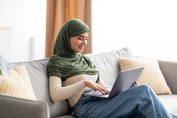 Portrait of young Arab female in hijab sitting on sofa, using laptop, surfing web, working or...
