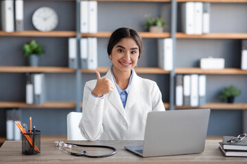 Happy friendly millennial pretty indian female doctor in coat works at laptop and shows thumb up