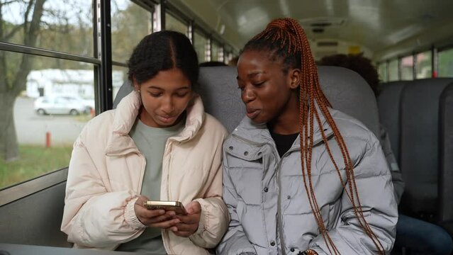 Smiling african american and indian schoolgirls watching interesting social media content on smartphone while riding school bus. Joyful multi-ethnic girls spending free time together after study