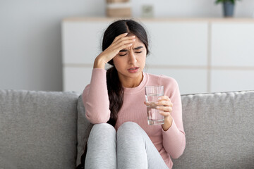 Despaired upset young indian lady suffer from fever and headache on sofa with glass of water in living room