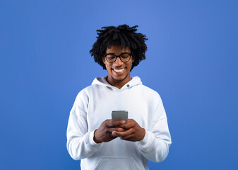 Happy black teenager looking at mobile phone screen, using new cool application on blue studio...
