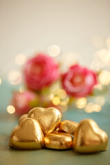 Heart shaped chocolates in golden foil. Blurred background with red flowers and yellow bokeh. Wedding greeting glamor card.