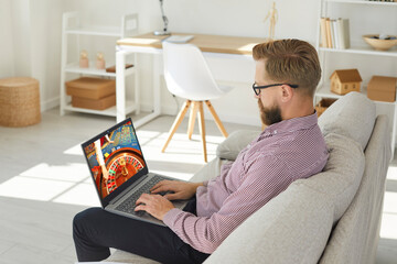 Young man sit relax on sofa play casino online on computer. Caucasian male rest at home couch...