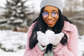 Young African American woman in glasses and warm gloves playing in the snow and spending time in...