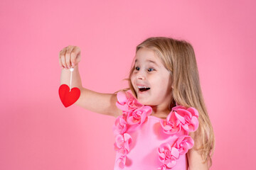 Funny little blue-eyed girl with long fair hair in chic pink dress stand smiling looking at red...