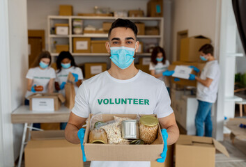 Charity center. Arab activist male volunteer holding food donation box and looking at camera, wearing medical mask