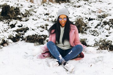 Young African American woman in glasses and warm gloves playing in the snow and spending time in the park outdoors