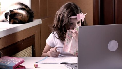 A little sick girl schoolgirl in a white T-shirt sits at a table with a laptop blows her nose into a handkerchief. Distance online learning for sick children. Meeting with friends via video call. - Powered by Adobe