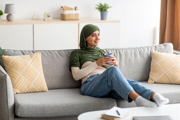 Obraz premium Pretty young Arab lady in hijab sitting on sofa with hot drink, spending peaceful weekend at home, copy space