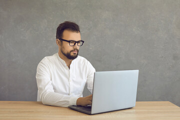Young businessman working on laptop sitting at desk against grey background. Professional male worker, recruiter manager or student engaged in remote work or training courses education e-learning