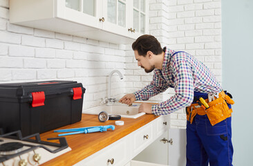 Obraz premium Professional plumber installs or repairs faucet and sink in kitchen of apartment. Side view of young skilled handyman in work wear who uses various tools to mount kitchen faucet in client's home.