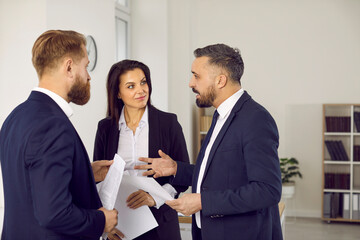Business people discuss contract to work together during meeting in modern office. Two men and woman in business suits stand with documents in hand and talk. Teamwork and partnership concept.